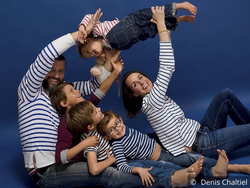 Photo de famille sur fond bleu, 2 parents et 4 enfants sourient, tous portent une marinière et un jean bleu.